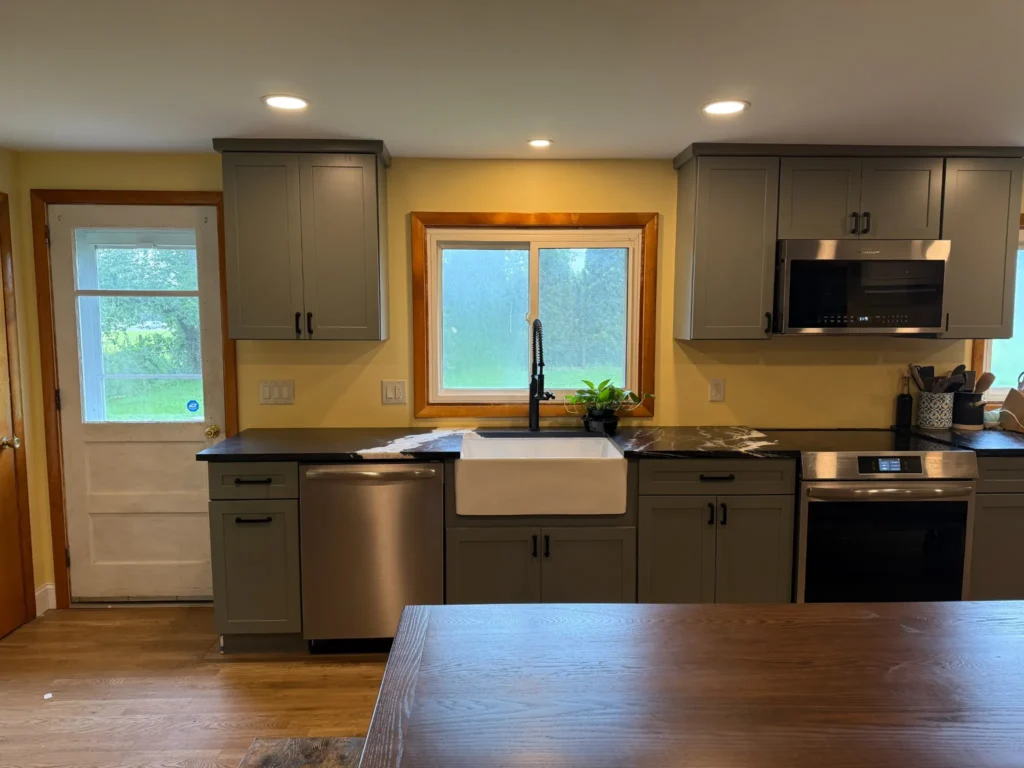 New sink wall with gray cabinets, a white farmhouse sink under the window, black countertops and stainless appliances